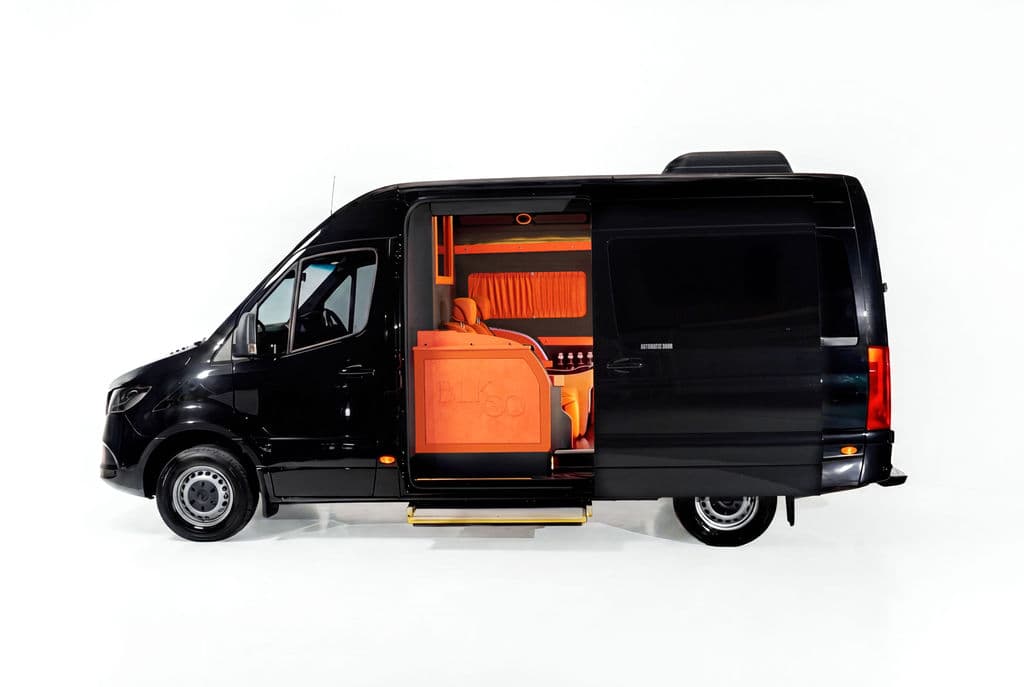 Black Mercedes-Benz Sprinter with the side door open on a tan captain-chair cabin, against a white studio sweep